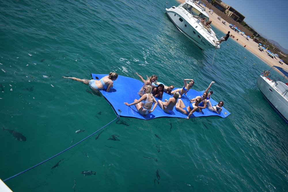 Group of friends lounging on a floating water mat beside an anchored yacht