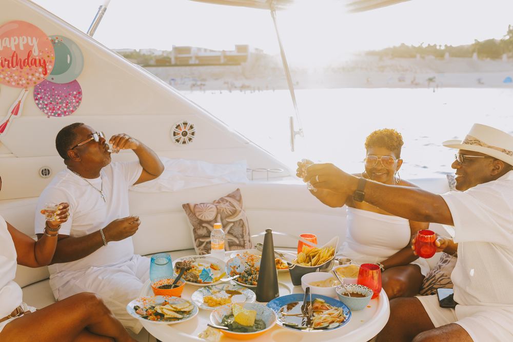 Friends in white outfits laughing and sharing food at a birthday celebration on a yacht deck